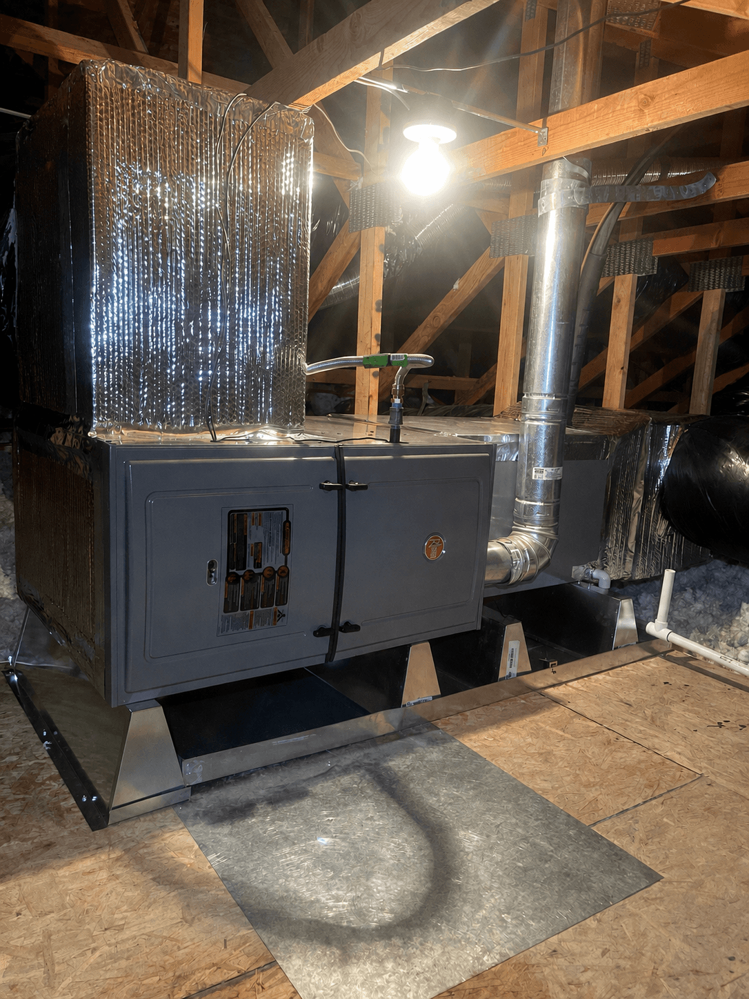 Gray HVAC unit with foil-wrapped ducts and metal piping installed in a wooden attic.