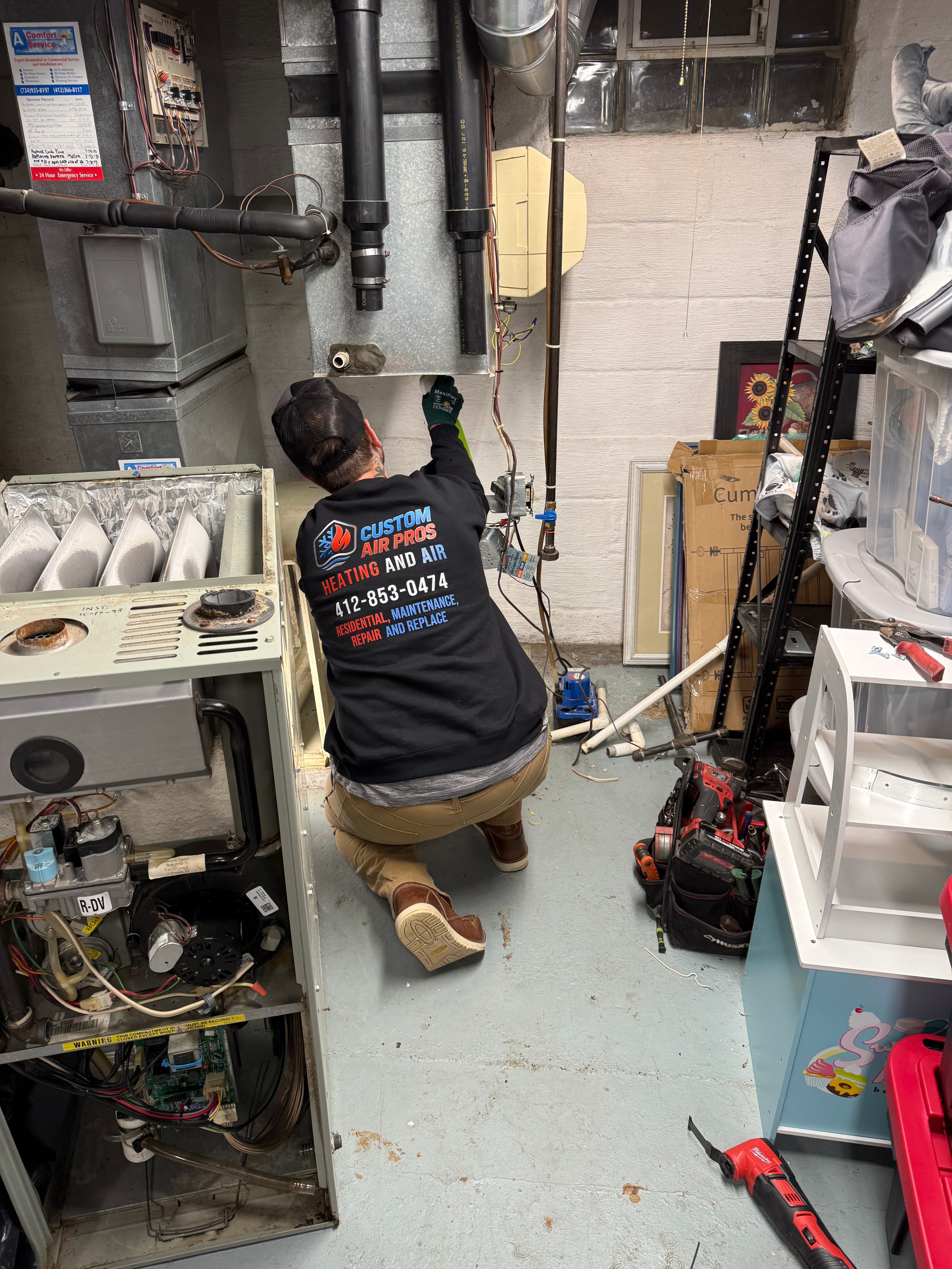 HVAC technician in a branded hoodie repairs a furnace in a basement with tools.