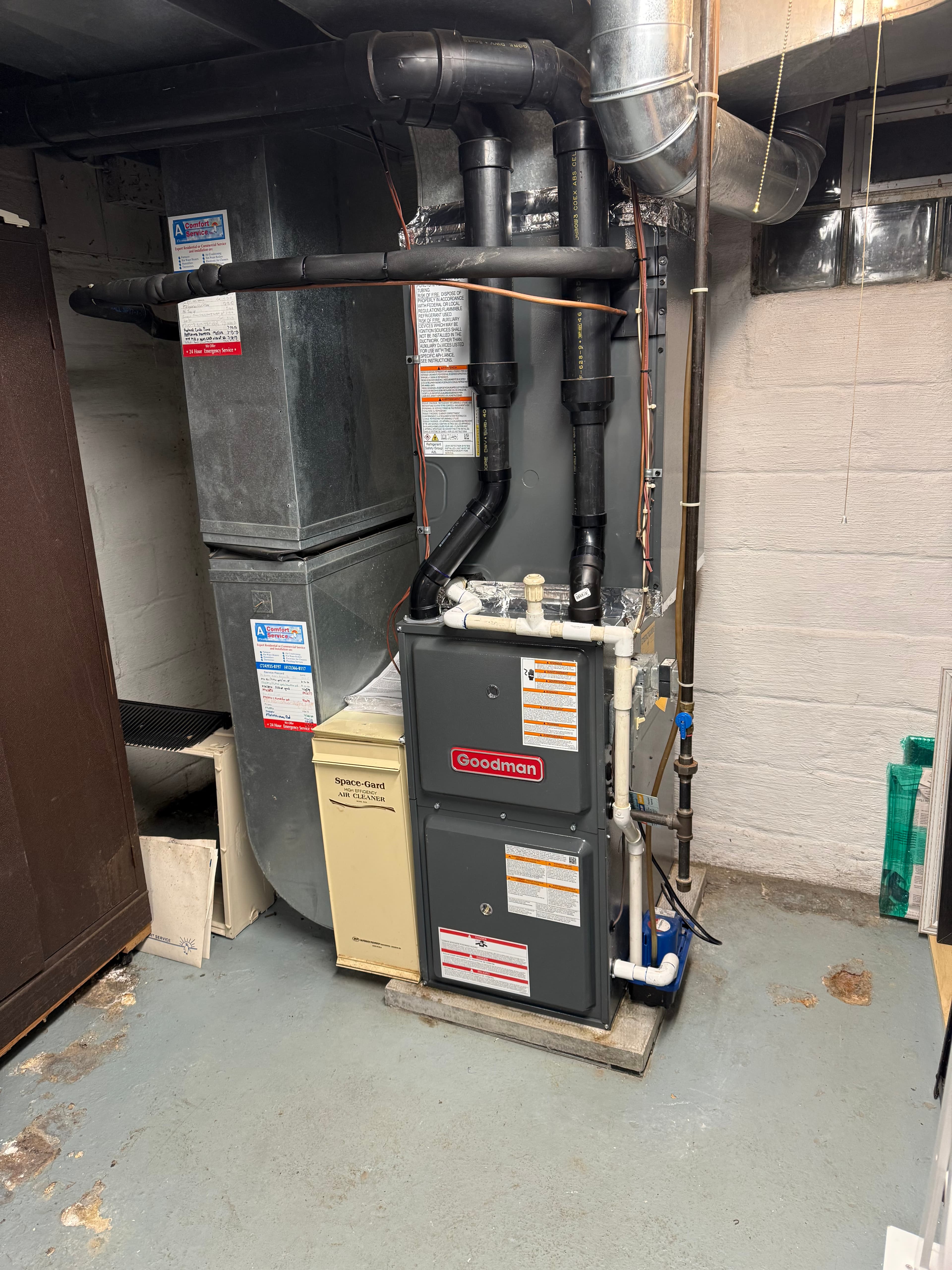 Gray Goodman furnace and beige air cleaner with black and white pipes in a basement.
