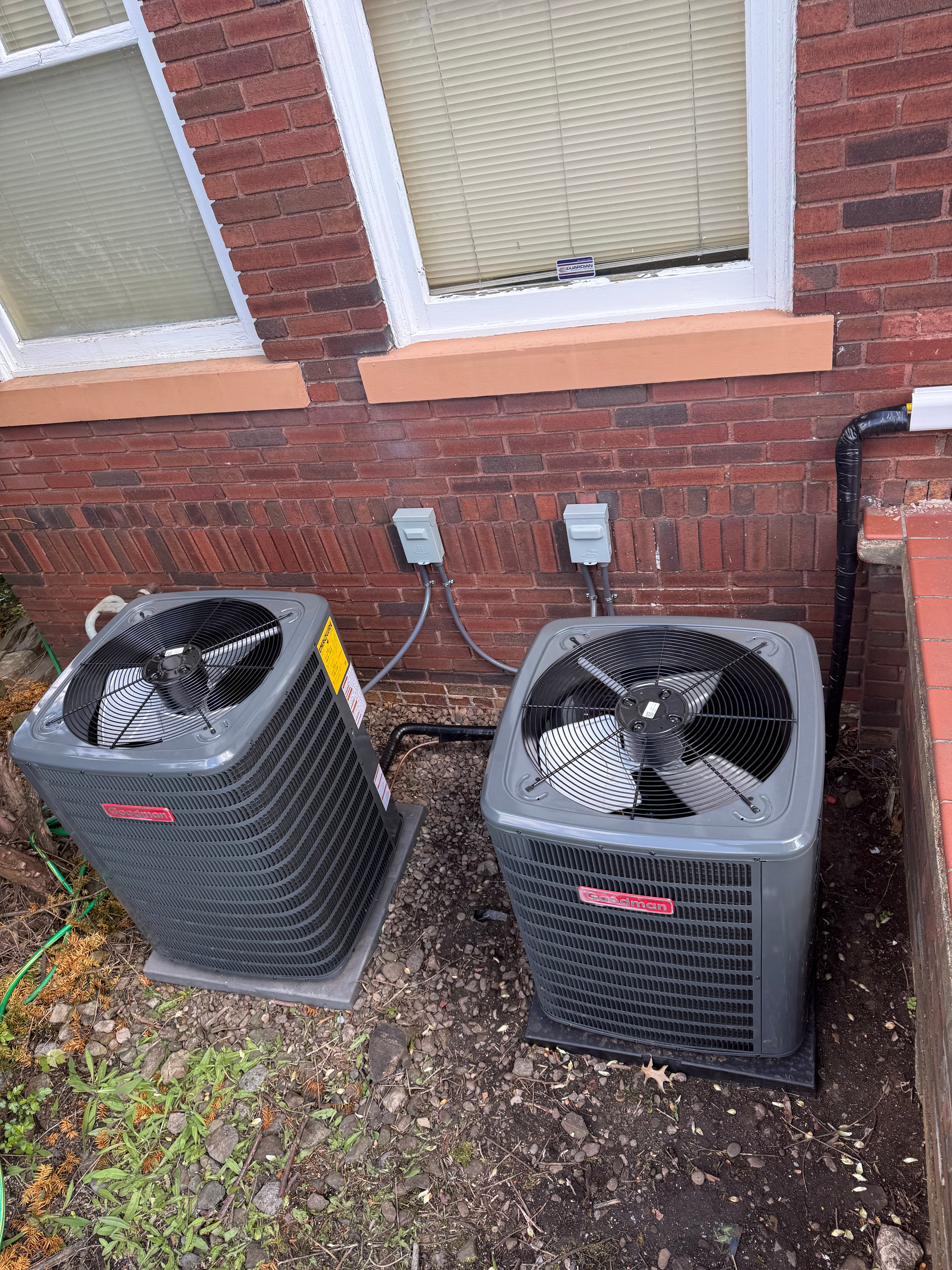Two gray Goodman air conditioning units installed outdoors against a red brick building wall.