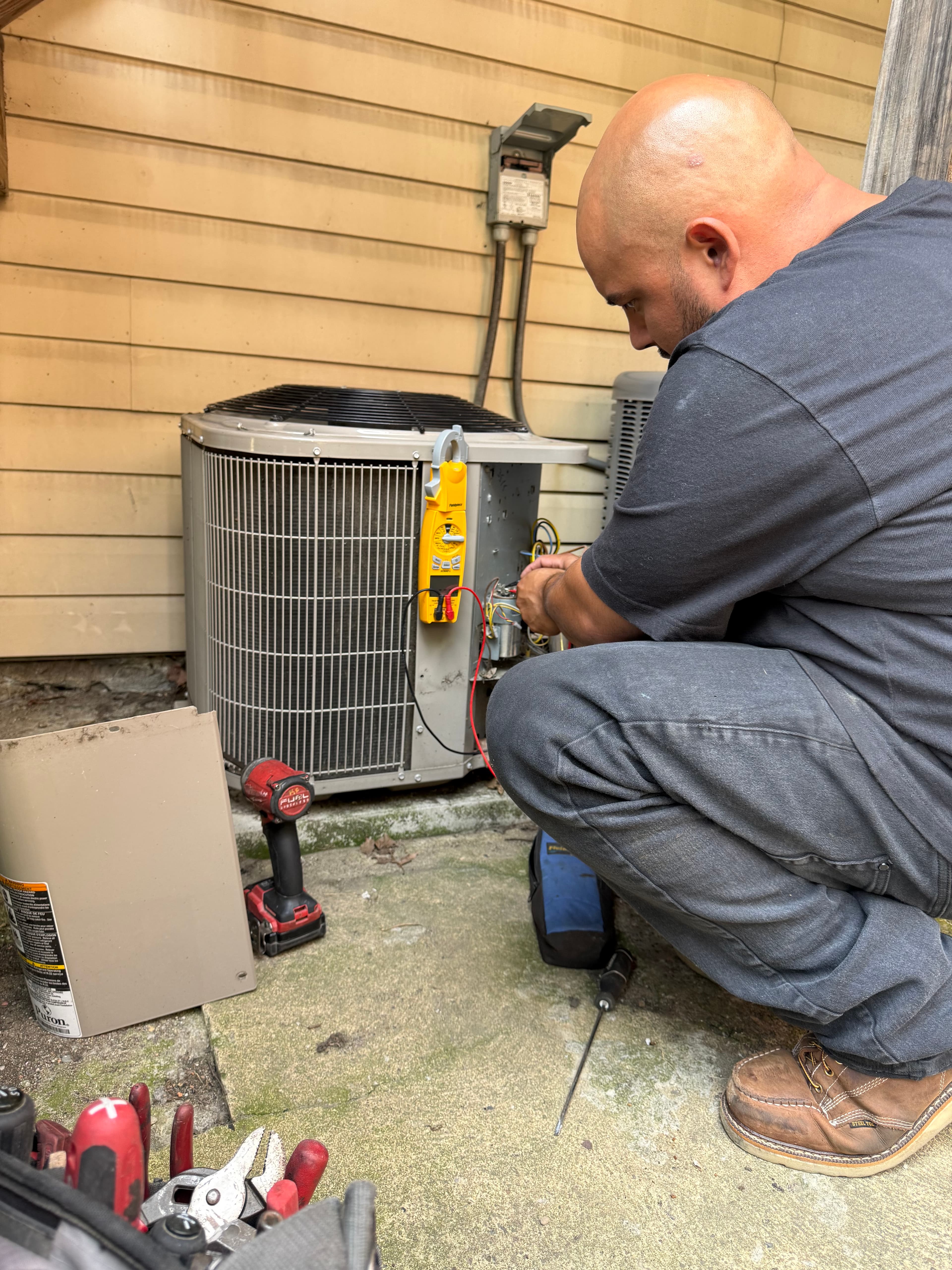 An HVAC technician repairs an outdoor air conditioning unit using a multimeter and tools.
