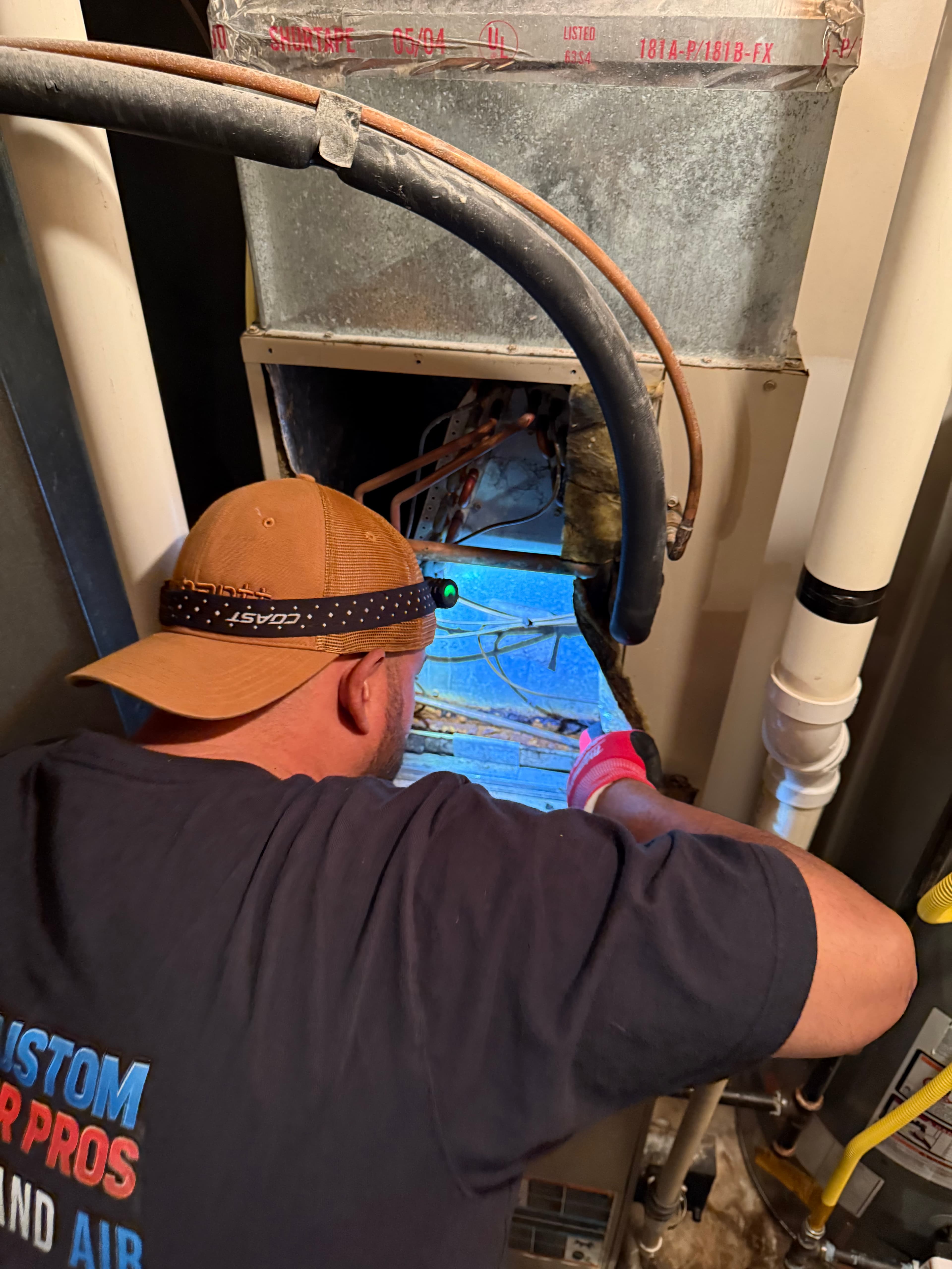 Technician with a headlamp inspects the blue-lit interior of an open HVAC furnace unit.