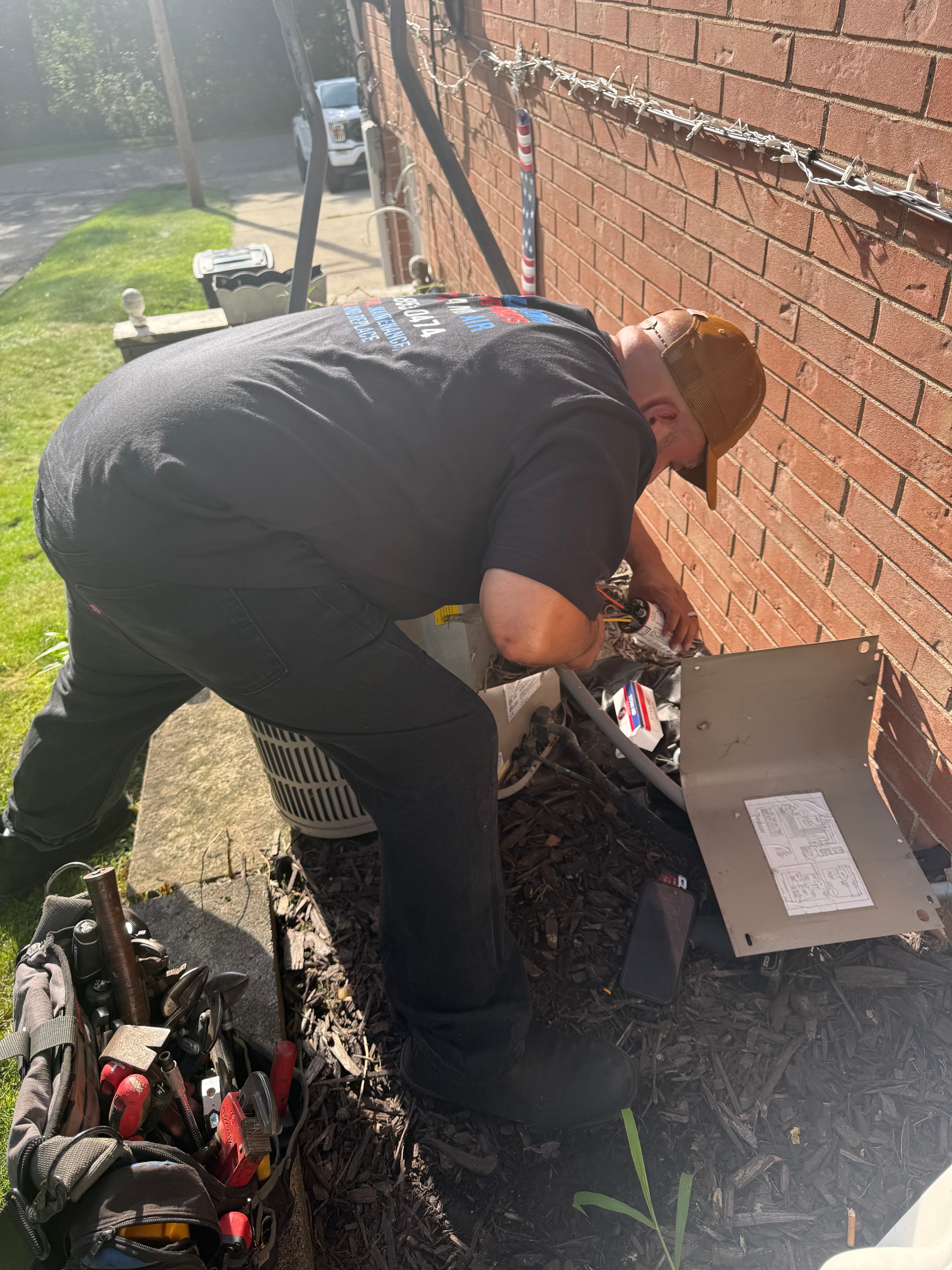 Technician in a black shirt repairs an outdoor AC unit next to a brick wall.
