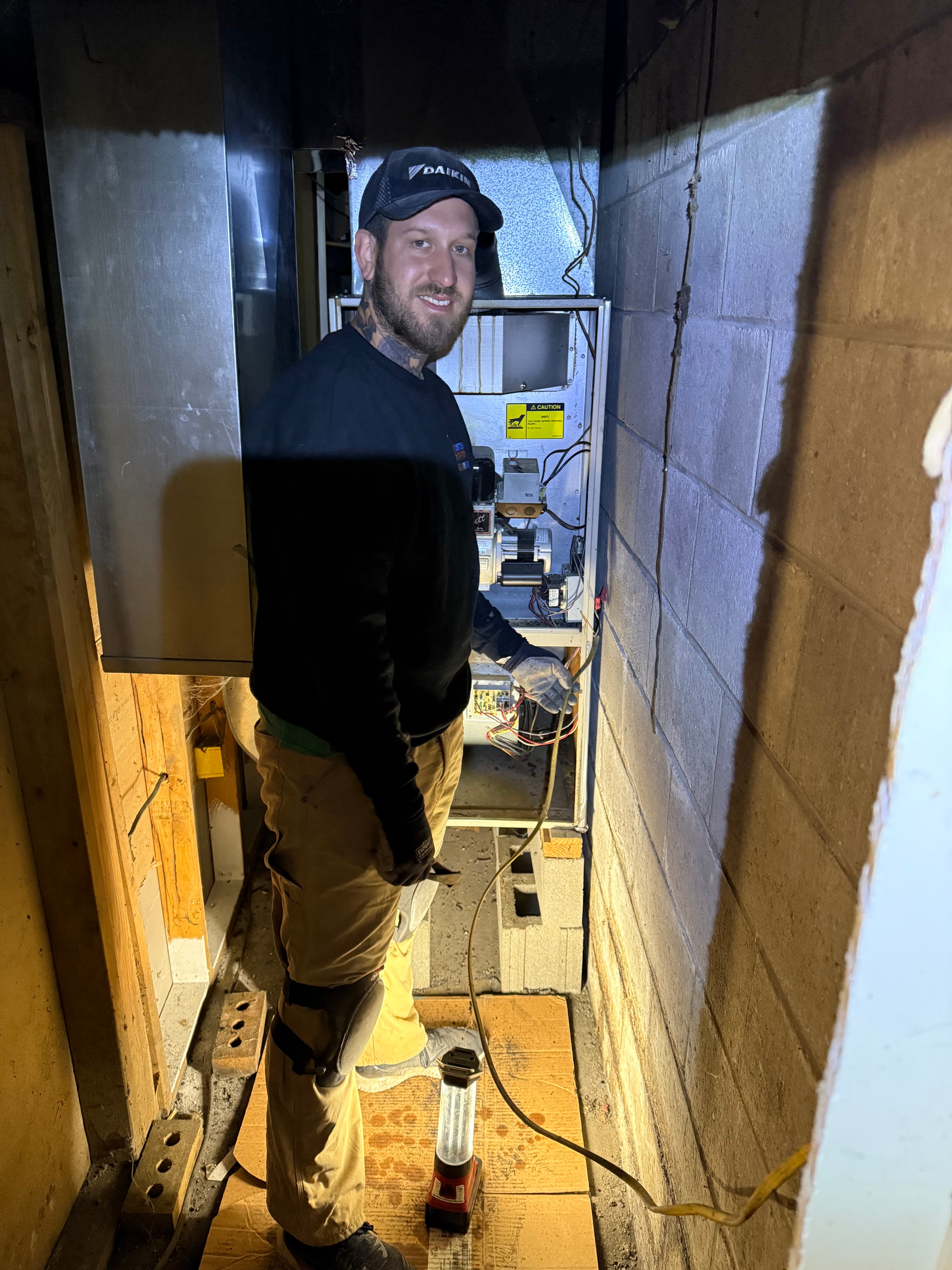 Smiling HVAC technician in a Daikin hat repairs an open furnace in a dark basement.
